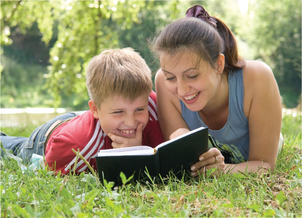 A photograph of a woman and a young boy reading a book together in a park. 